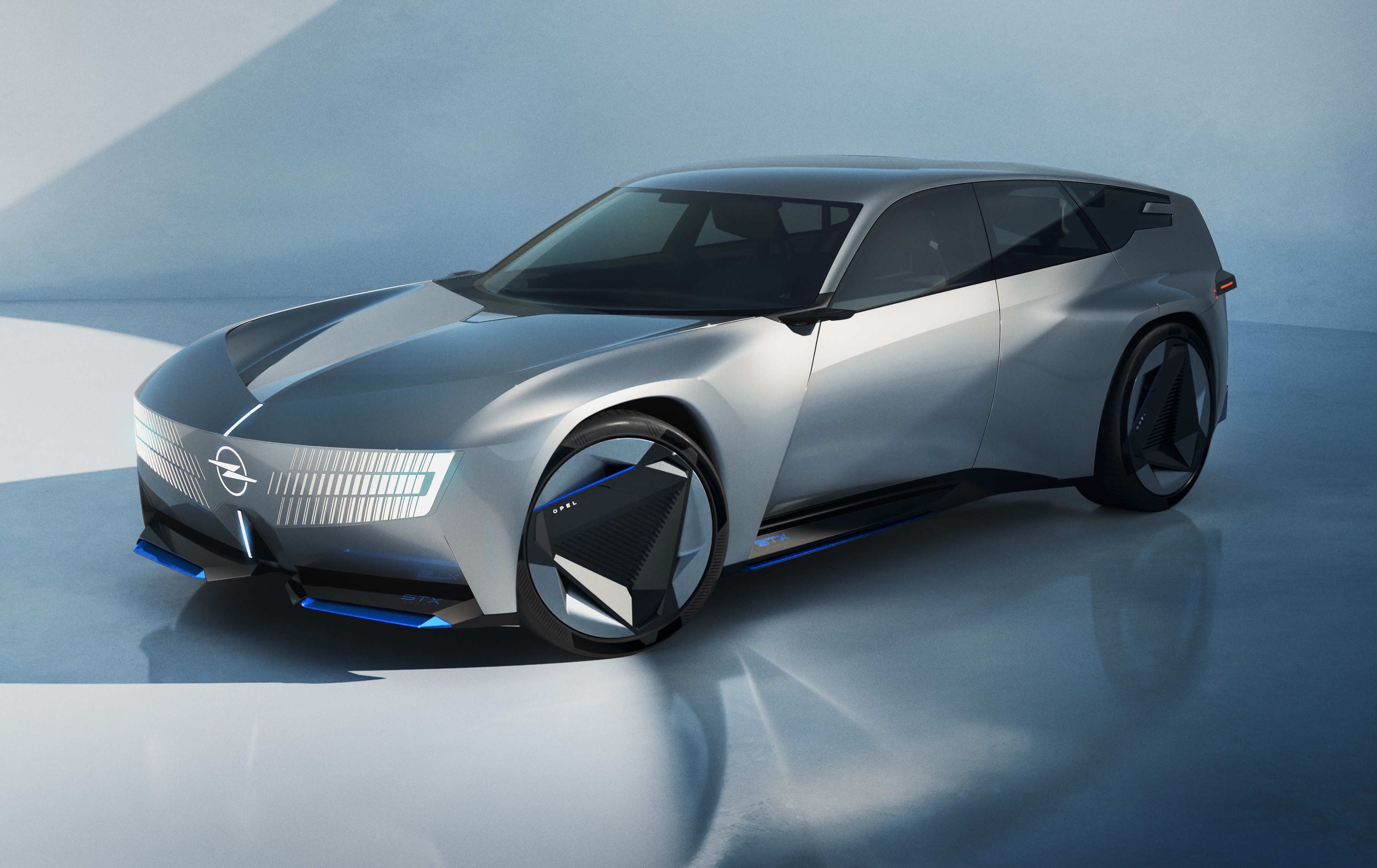 Carro conceito Opel Experimental prateado, com design futurista, linhas aerodinâmicas e detalhes azuis na parte inferior e nas rodas. O veículo está parado em um ambiente minimalista com paredes e chão claros, refletindo a luz. O logotipo da Opel está na grade frontal, que possui iluminação horizontal em LED