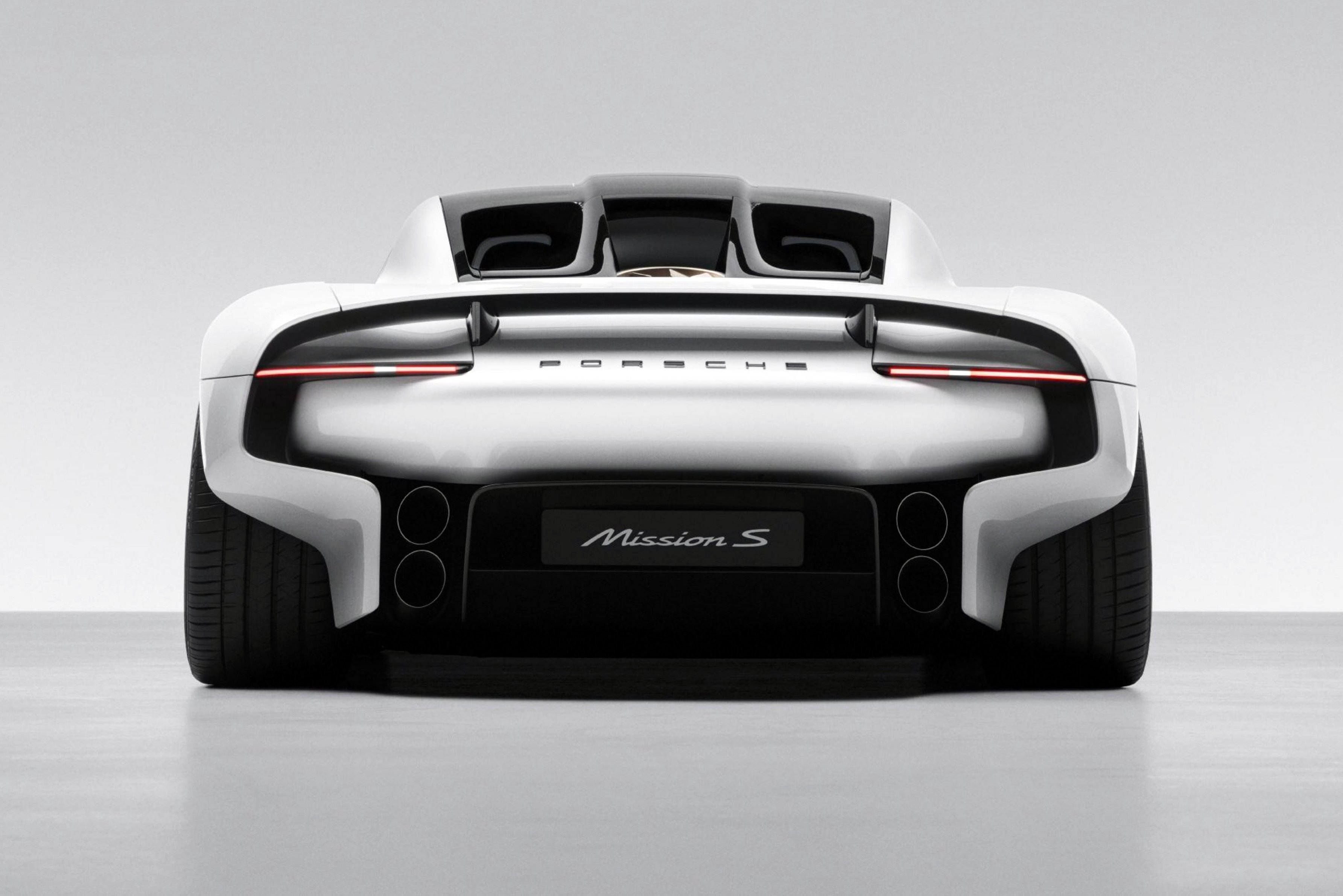 Traseira de um carro esportivo branco, modelo Porsche Mission S, com lanternas horizontais vermelhas acesas, quatro saídas de escape pretas e o nome Porsche em relevo no porta-malas. O carro está em um estúdio com fundo cinza claro e chão liso
