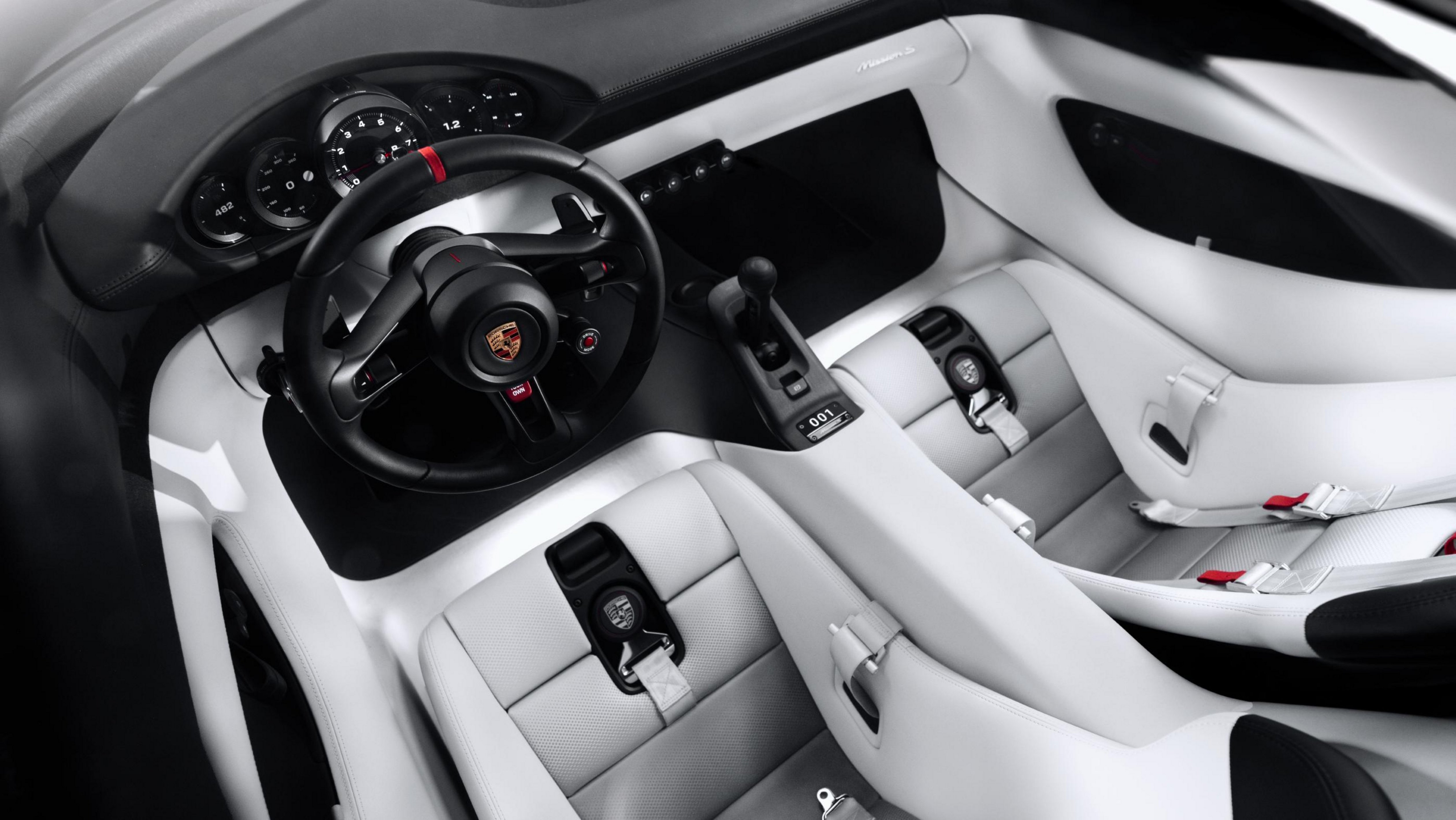 Interior de um carro esportivo Porsche, com volante preto e detalhes vermelhos, painel de instrumentos analógico, câmbio manual e bancos esportivos brancos com cintos de segurança de quatro pontos