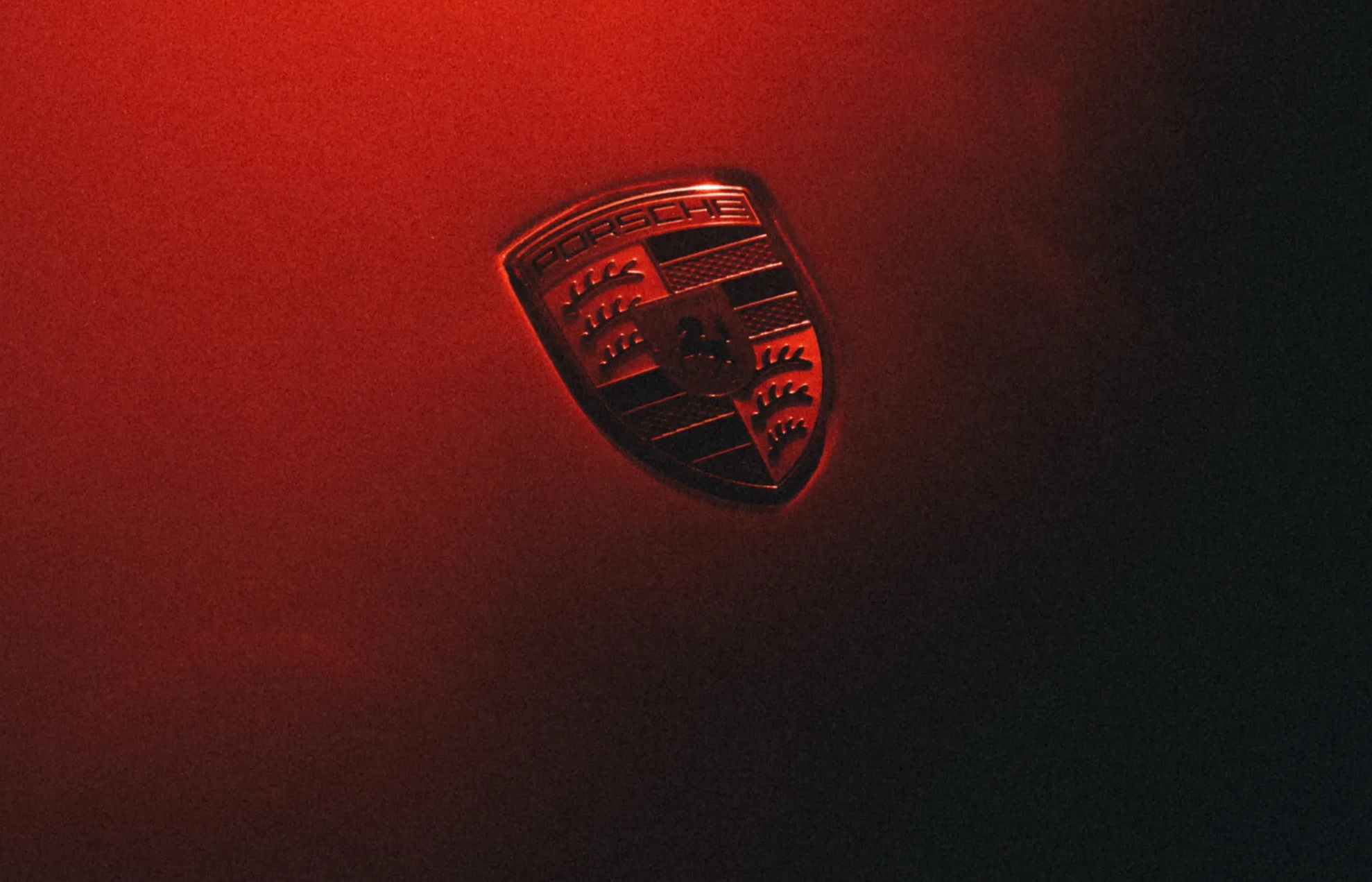 Emblema da Porsche em tons de vermelho e preto, com o nome da marca no topo e um cavalo rampante ao centro, sobre um fundo escuro