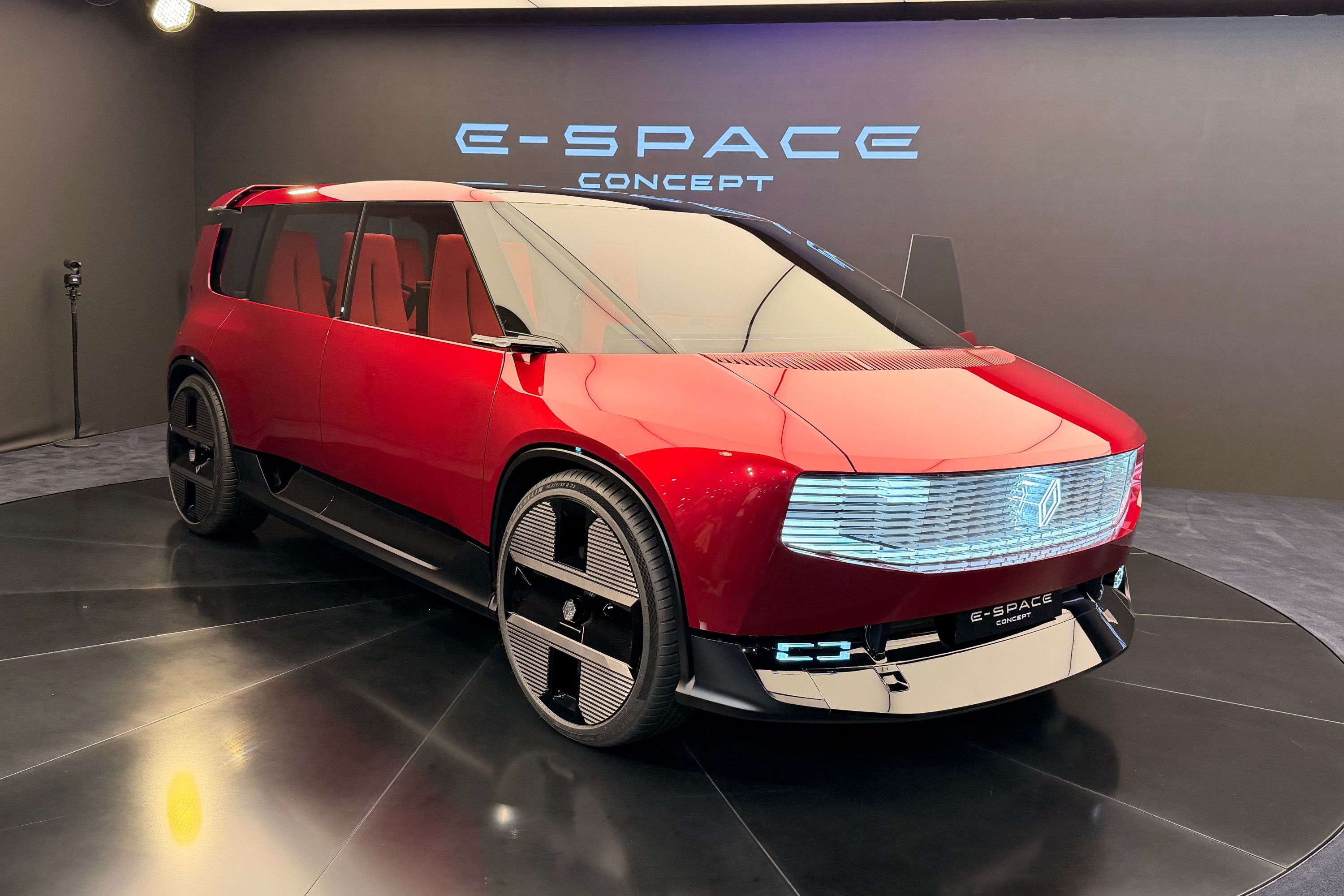 Carro conceito E-Space vermelho brilhante, com design futurista, faróis e grade iluminados por LED, rodas pretas com detalhes prateados, em um showroom escuro com o nome E-SPACE CONCEPT iluminado ao fundo