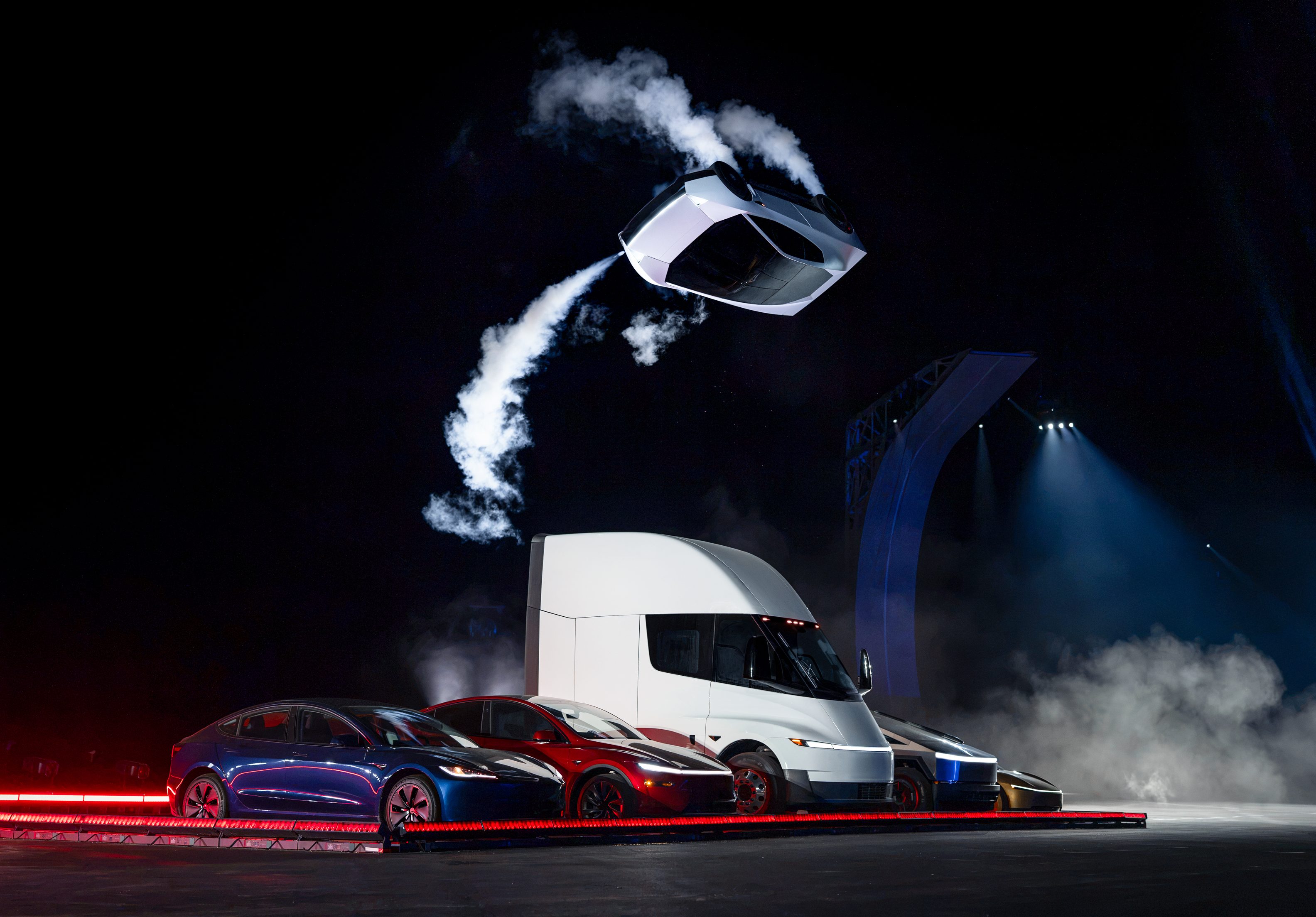 Caminhão branco Tesla Semi e dois carros esportivos, um azul e um vermelho, em uma plataforma iluminada por luzes vermelhas. Fumaça branca sobe do caminhão e de um objeto flutuante acima, em um palco escuro com luzes azuis ao fundo