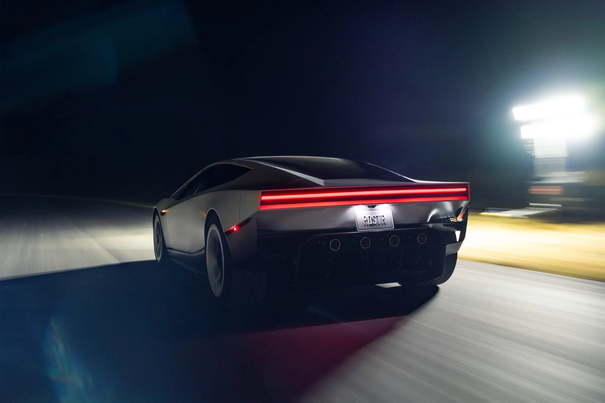 Carro esportivo cinza escuro visto por trás, com lanterna traseira vermelha horizontal e placa POSTR, em movimento rápido à noite, com luzes de farol iluminando a pista e postes de luz ao fundo