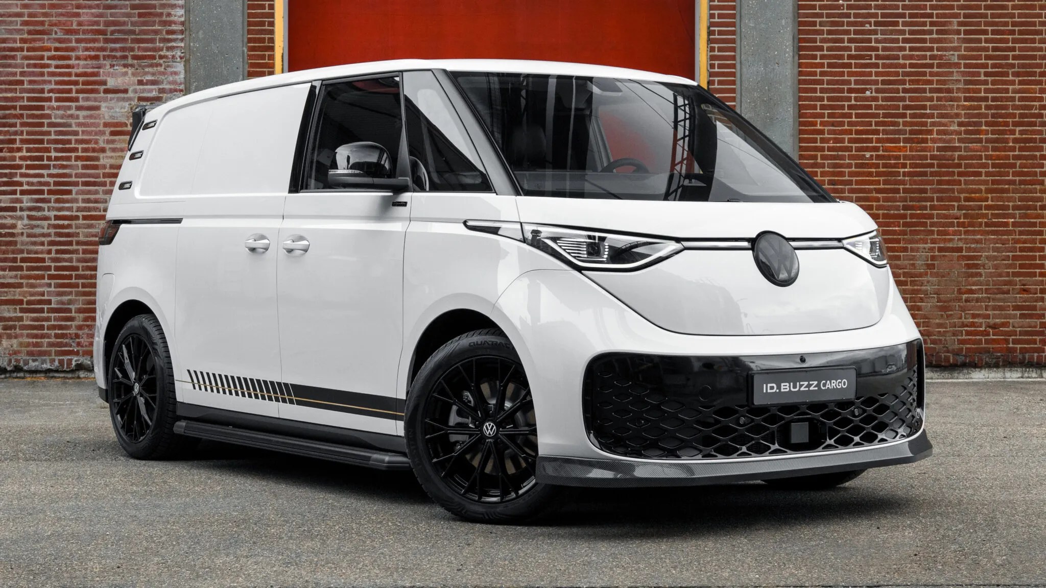 Van Volkswagen ID. Buzz Cargo branca, com detalhes esportivos pretos, rodas pretas e faixas decorativas laterais, estacionada em frente a uma parede de tijolos e uma porta vermelha