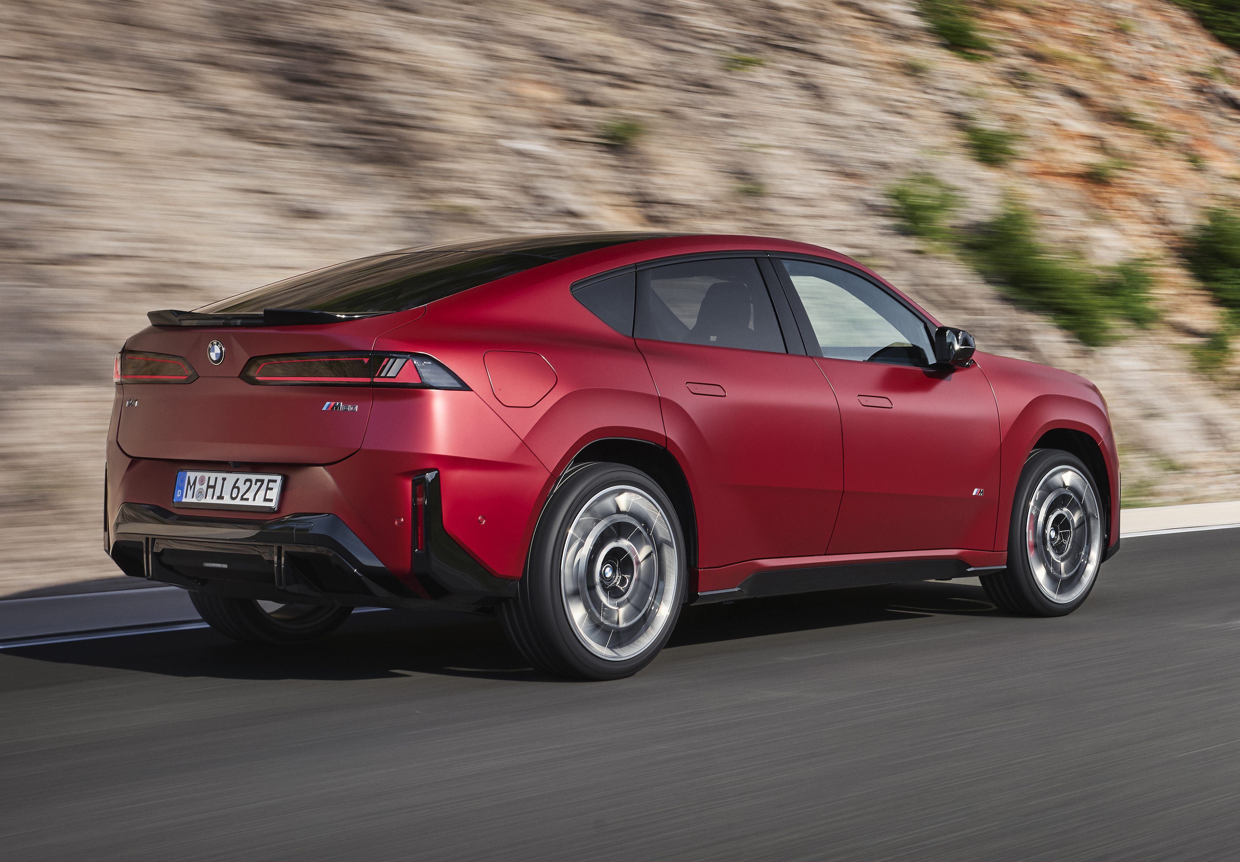 Carro vermelho escuro, modelo SUV cupê, em movimento rápido em uma estrada asfaltada. O veículo tem design moderno, com teto inclinado e rodas prateadas. Ao fundo, uma encosta de terra e vegetação borrada pelo movimento
