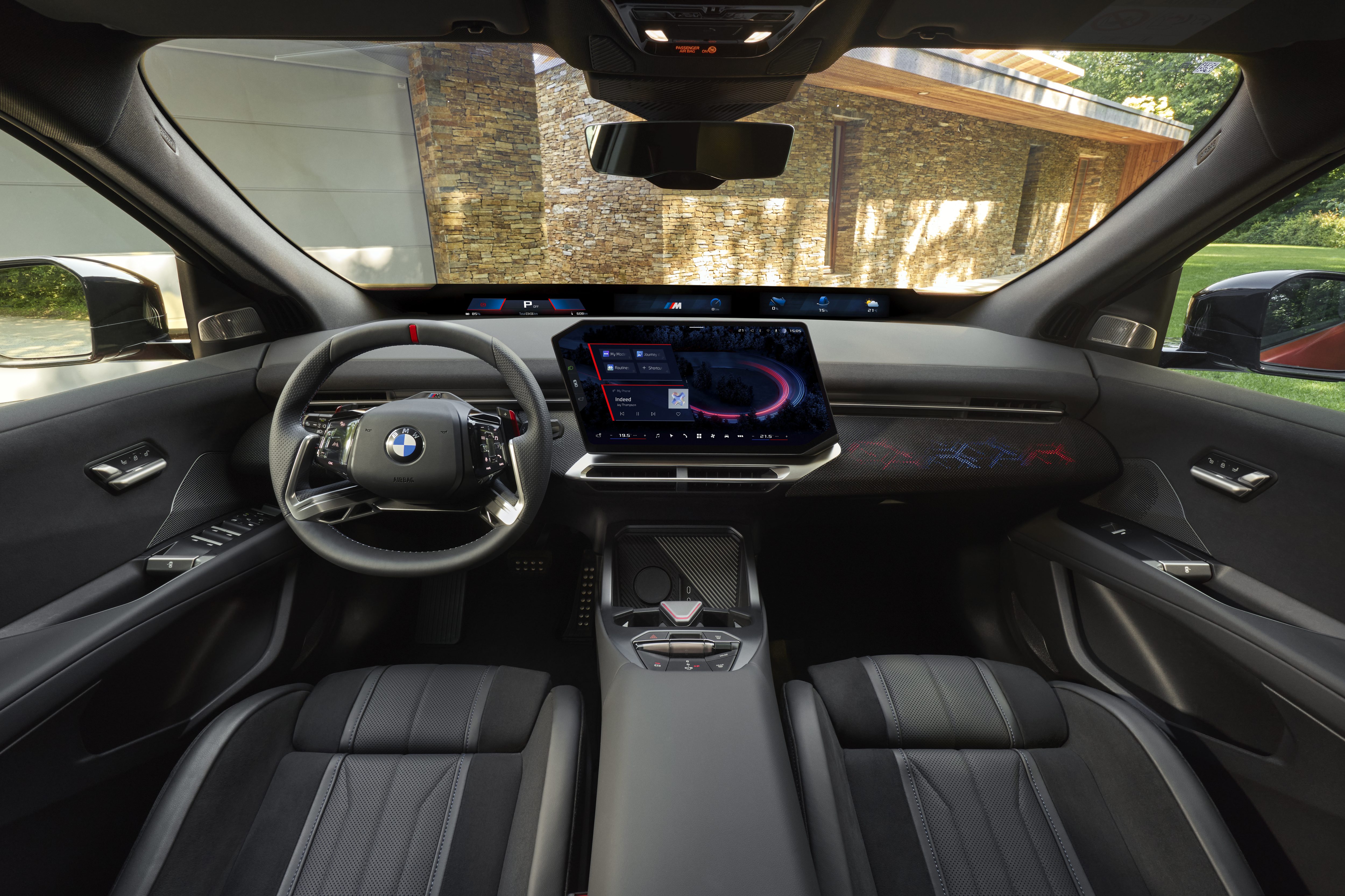 Interior de um carro BMW moderno, com volante esportivo e painel digital curvo exibindo informações de navegação e mídia. Os bancos são pretos com detalhes perfurados e costura azul. Ao fundo, uma casa com parede de pedra e vegetação verde é visível através do para-brisa