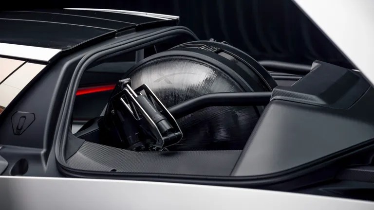 Um cilindro de gás natural veicular (GNV) preto e prateado, com uma válvula metálica, instalado no porta-malas de um carro cinza, com acabamento interno escuro e detalhes em fibra de carbono