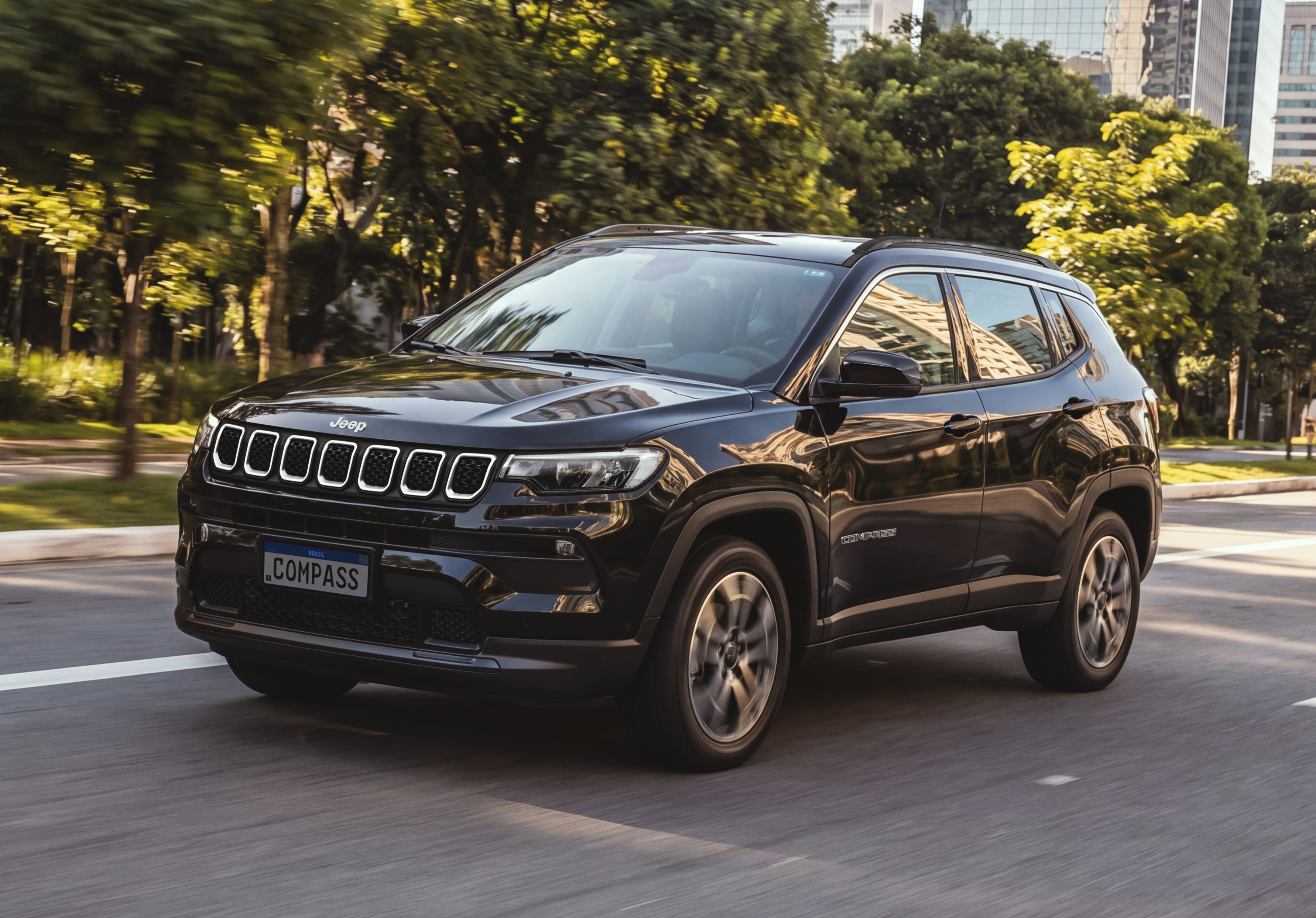 SUV Jeep Compass preto em movimento, visto de frente e lateral, em uma avenida arborizada com prédios ao fundo