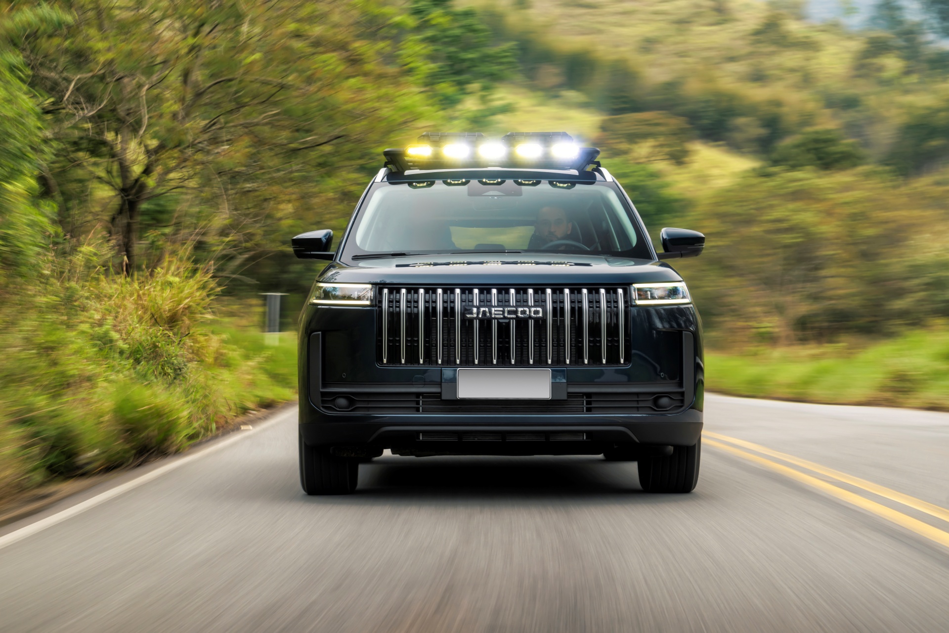 SUV preto Jaecoo com faróis auxiliares no teto, em movimento rápido por uma estrada asfaltada com faixa dupla amarela, cercada por vegetação verde e borrada pelo movimento
