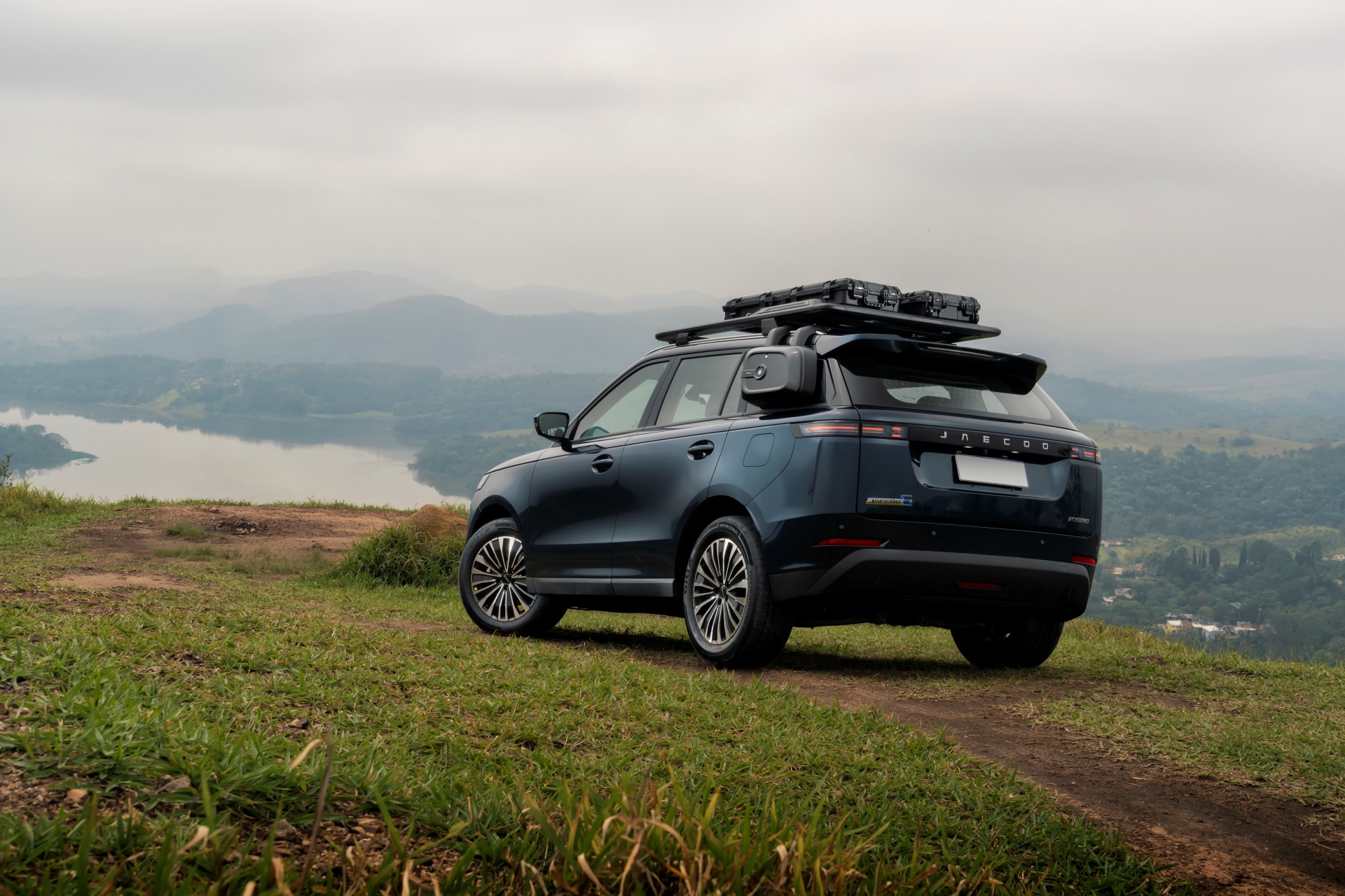 SUV azul-escuro com bagageiro de teto e caixas de armazenamento, estacionado em uma trilha de terra em um morro gramado, com um lago e montanhas ao fundo sob um céu nublado