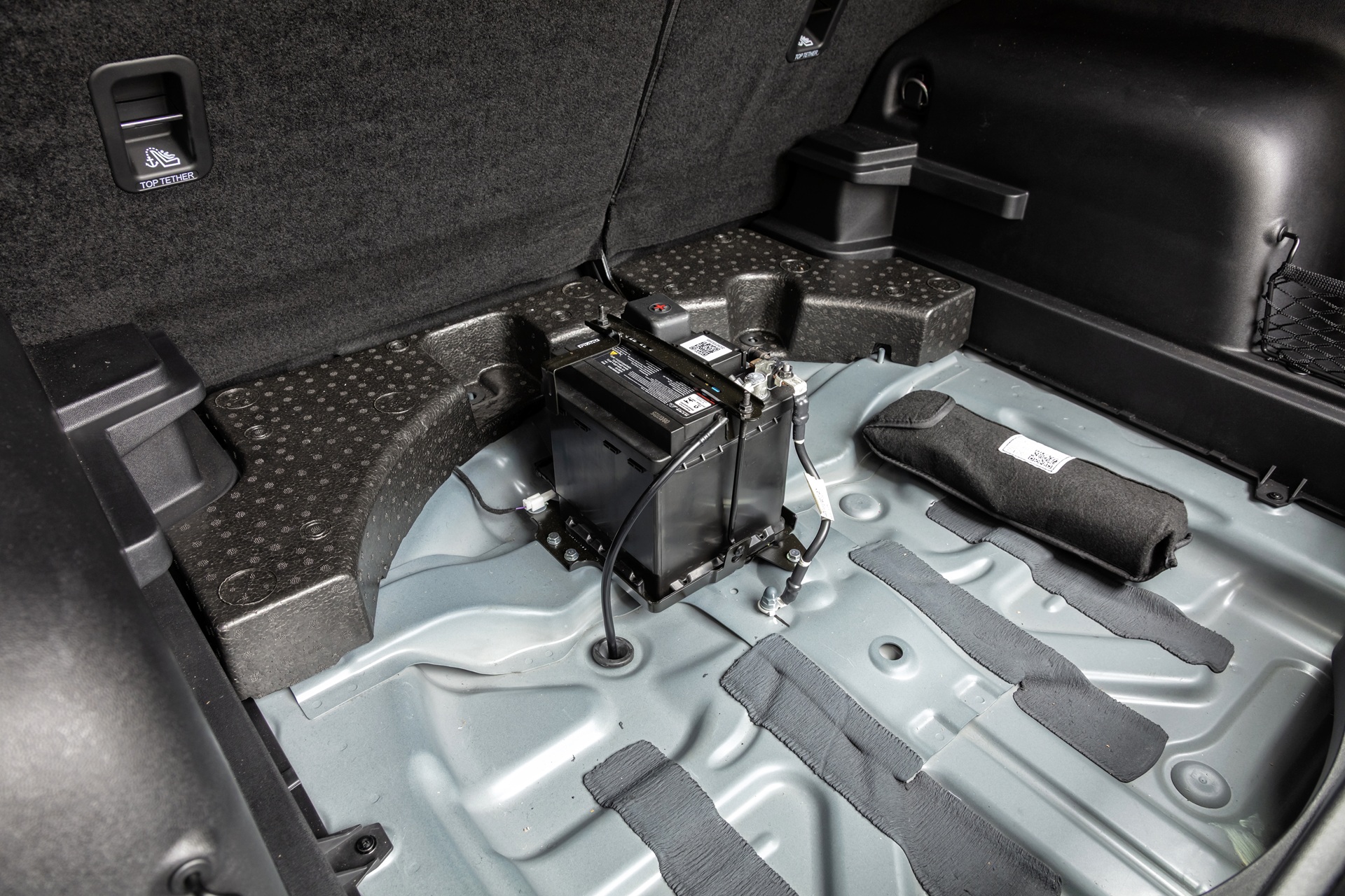 Compartimento de bagagem de um carro, com a bateria preta exposta no centro, cabos conectados e um kit de ferramentas enrolado ao lado, sobre o assoalho cinza metálico