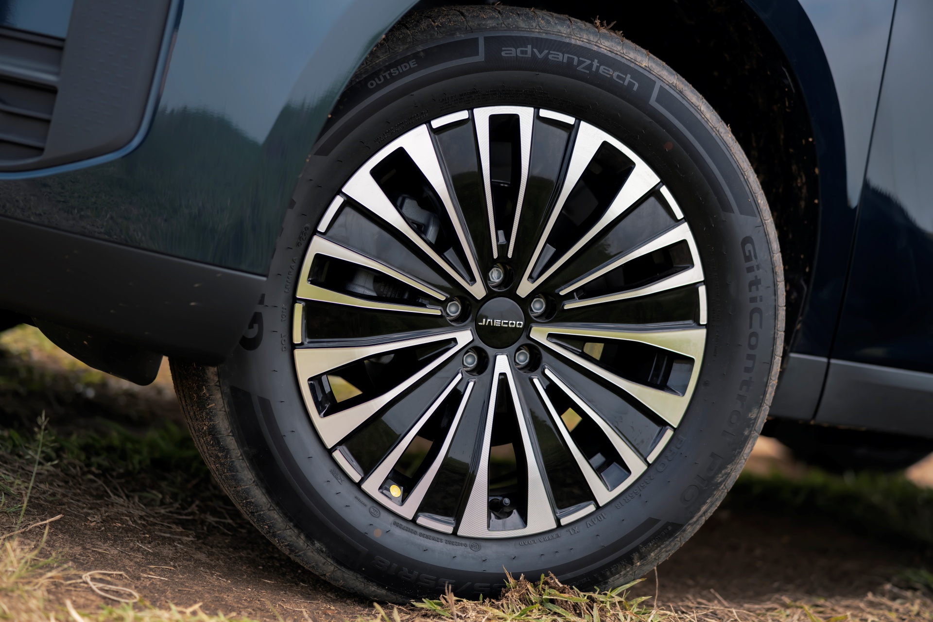Roda de carro com aro preto e raios prateados, pneu preto com inscrição Advanztech e Giticon, parte da lataria azul-escura do veículo visível acima, sobre chão de terra e grama seca