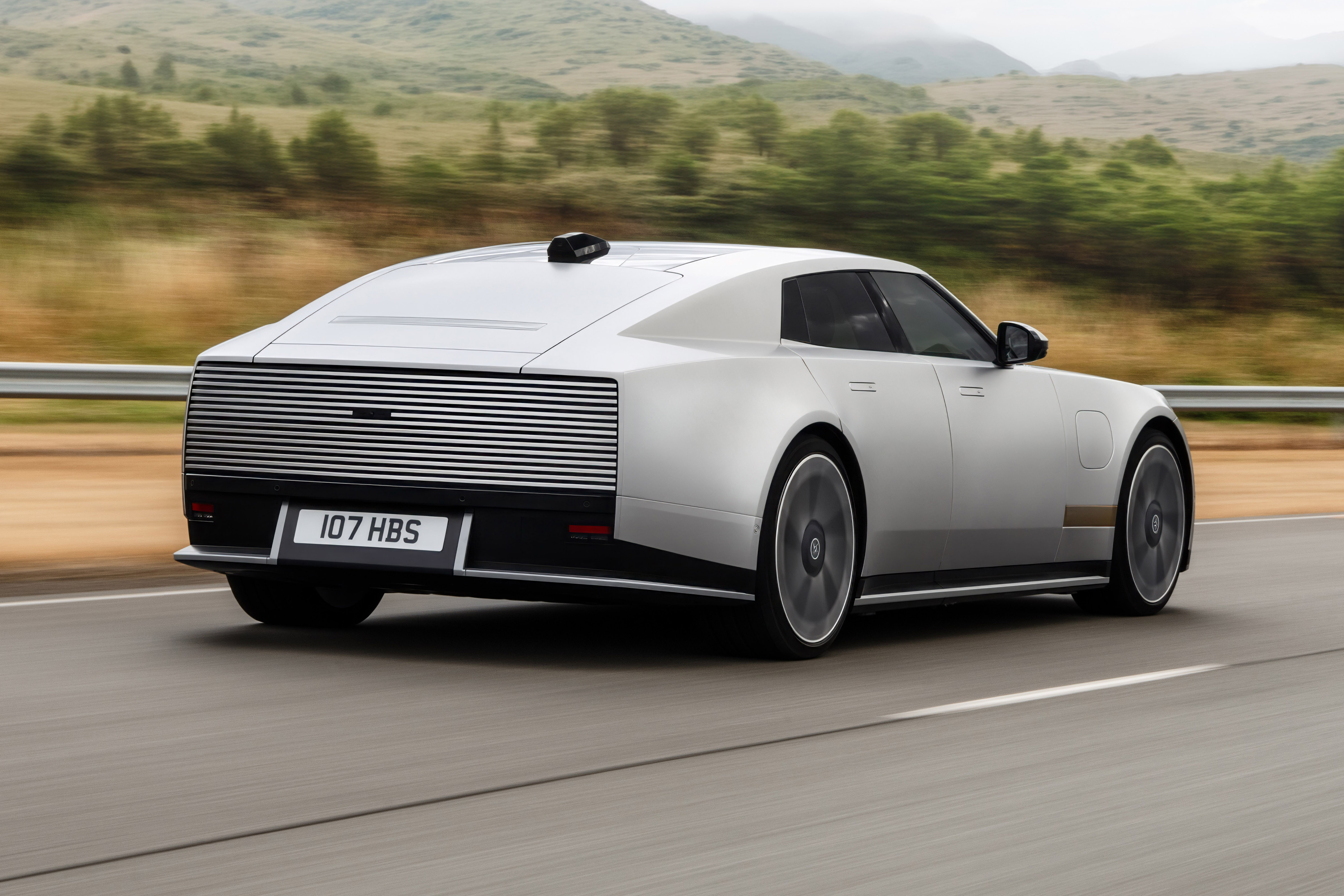 Carro prateado de design futurista, com traseira retangular e lanternas horizontais, em movimento rápido numa estrada com montanhas e vegetação ao fundo