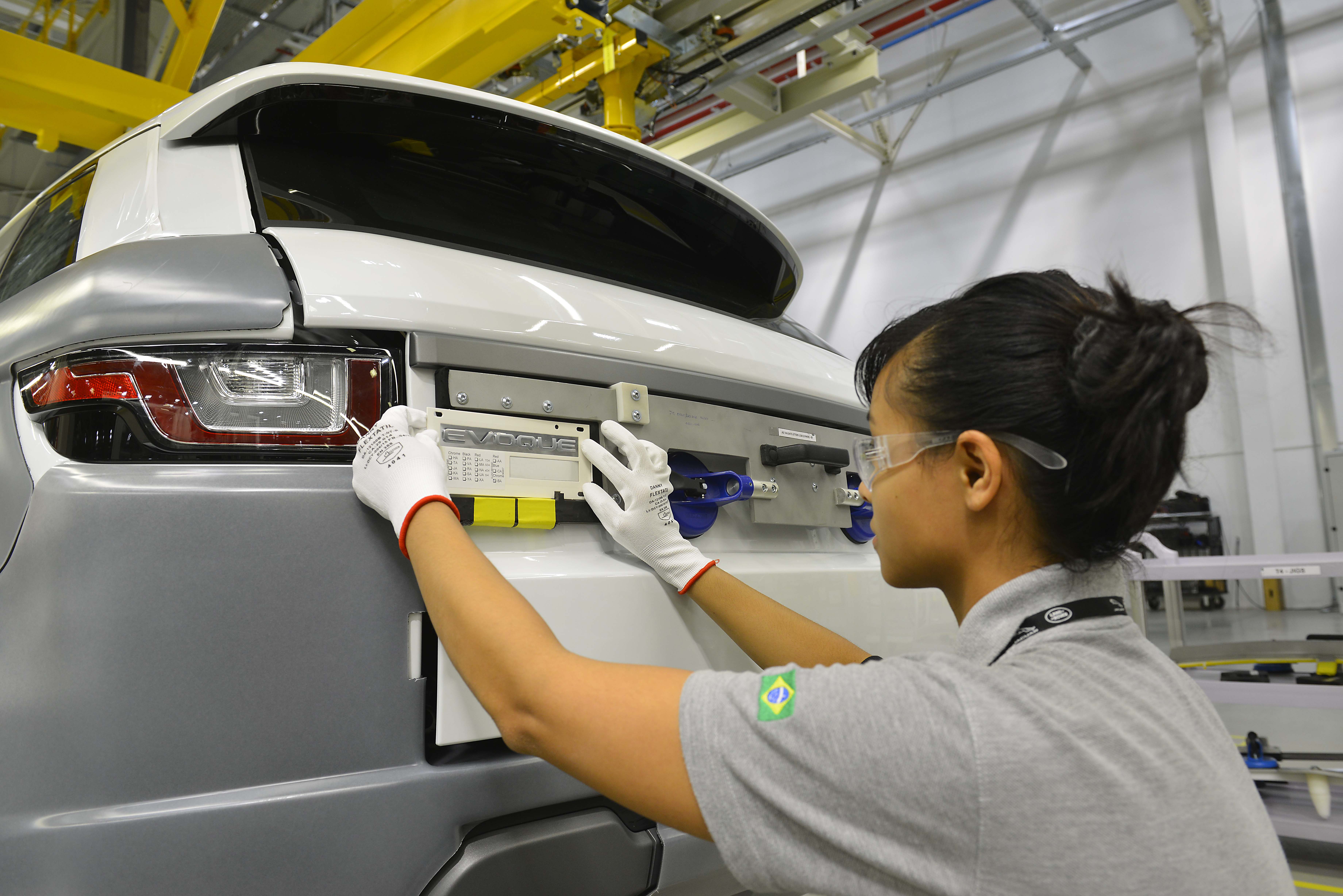 Mulher de óculos de proteção e luvas brancas com borda vermelha, montando a traseira de um carro branco e cinza em uma fábrica. Ela usa uma camiseta cinza com a bandeira do Brasil no braço direito e tem o cabelo preso em um coque. O carro tem a palavra EVOQUE visível na parte traseira.