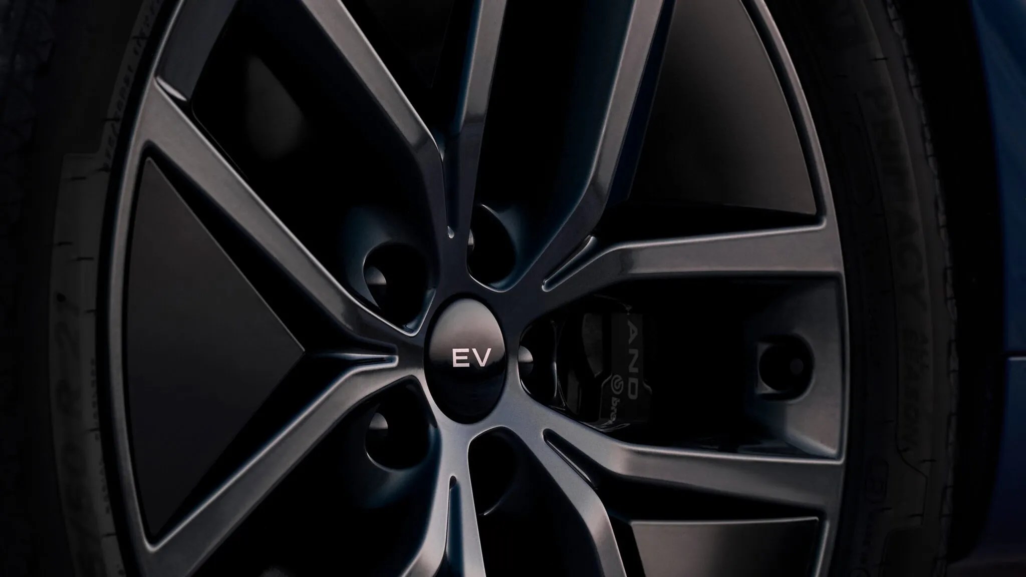 Roda de carro com design moderno em tons de cinza escuro e preto, pneu lateral visível. O centro da roda tem um emblema redondo com as letras EV em branco, indicando um veículo elétrico